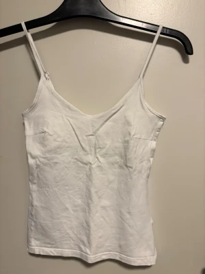 Vit linnetopp med smala axelband - Säljer en enkel och stilren vit linnetopp med smala justerbara axelband. Toppen har en v-ringad hals och är figurnära i modellen. Perfekt för varma dagar och lätt att matcha med olika outfits. Materialet känns mjukt och bekvämt mot huden.