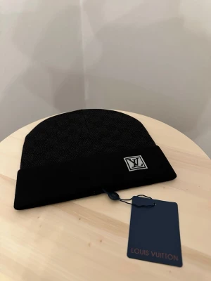 Svart L.V mössa med logga - Snygg svart mössa från L.V med diskret rutmönster och uppvikt kant. Märkets klassiska L.V-logga sitter framtill på en patch. Mössan är tillverkad i ett mjukt och värmande material som passar perfekt för kyliga dagar.