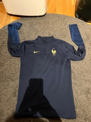 Nike Dri-Fit FFF träningströja marinblå - Säljer en marinblå Nike Dri-Fit träningströja med guldiga detaljer och FFF-logga på bröstet. Tröjan har halv dragkedja, slim fit-passning och mönstrade ärmar i blått. Perfekt för fotbollsträning och sportiga aktiviteter.