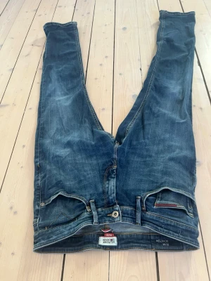 Blå Hilfiger Denim jeans slim fit - Säljer ett par blå jeans från Hilfiger Denim i slim fit-modell. Jeansen har klassisk femficksdesign, snygga slitningar och detaljer med Hilfiger-logga på bakfickan och läderpatch i midjan. Tillverkade i slitstarkt jeansmaterial med gul kontrastsöm. Ny pris 1200 mitt pris 225kr