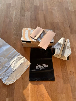 Golden Goose vita sneakers med stjärna - Golden Goose sneakers i vitt skinn med grå mockadetaljer och ikonisk stjärna på sidan. Platt sula och rund tå, snörning framtill och svart hälparti med logotyp. Kommer med originalkartong, dustbag och pass. Perfekt för dig som gillar coola och trendiga sneakers.