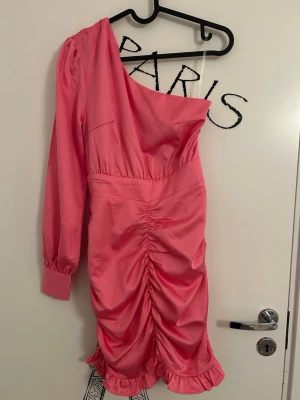 Rosa oneshoulder klänning från Glamorous - Rosa oneshoulder klänning från Glamorous i storlek 34. Klänningen har lång ärm med knappar vid manschetten, rynkade detaljer längs framsidan och volangkant nedtill. Väldigt fin klänning som är i nytt skick.
