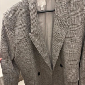 Rutig kavaj från H&M - Snygg rutig kavaj från H&M i klassisk grå färg med svarta knappar. Kavajen har en rak passform, breda slag och är helfodrad. Perfekt för dig som vill ha en stilren och trendig look med lite edge.