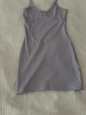 Ljuslila randig klänning  från H&M - Säljer en ljuslila klänning från H&M Divided i storlek S. Toppen har tunna axelband, ribbat tyg med smala ränder, spetsdetaljer längs halsringningen och en liten rosett framtill. Perfekt för sommarens varma dagar.
