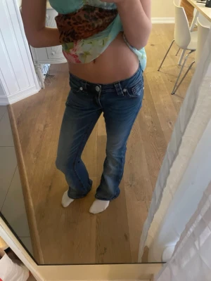 Blå bootcut lågmidjade jeans - Säljer ett par blå jeans med bootcut och låg midja. Jeansen har klassisk femficksdesign, snygga slitningar och en lätt tvättad look. Materialet är jeans och passformen är normal med lite utsvängda ben. Perfekta för dig som gillar retrovibbar och vill ha en avslappnad stil. I storlek XS young 14+ använda ett par gånger