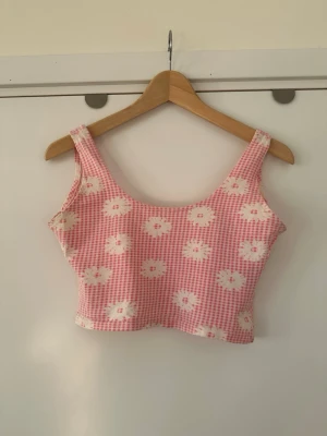 Rutig rosa croppat linne - Superfin croppad topp med rosa och vita rutor och blommigt mönster. Toppen har breda axelband och är ärmlös, perfekt för varma dagar. Materialet känns mjukt och lätt, och den har en söt, ungdomlig vibe.