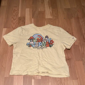 Gul t-shirt från Tommy Jeans S - Gul t-shirt från Tommy Jeans med färgglatt tryck framtill med svamp, blommor och regnbåge. Skön passform och klassisk rund hals. Perfekt för dig som gillar retro vibes och coola prints.