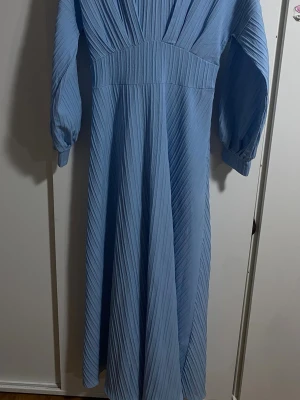 Ljusblå långklänning med plissering - Säljer en ljusblå långklänning med plisserat tyg och långärmade ärmar. Klänningen har en markerad midja och ett snyggt fall. Stängs med dragkedja i ryggen och har en elegant, stilren look som passar perfekt till vår och sommar. Den snatchar midjan perfekt