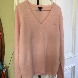  GANT V-ringat tröja i superfin lammull. - Superfin ljusrosa stickad tröja från GANT med v-ringning och broderad logga på bröstet. Tröjan har långa ärmar och ribbade muddar vid ärmslut och nederkant. Perfekt för dig som gillar en klassisk och chill stil.