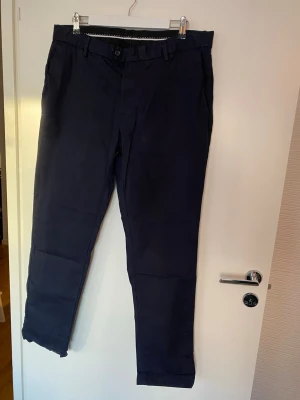 Mörkblå kostymbyxor från Bläck - Snygga mörkblå kostymbyxor från Bläck i klassisk rak modell. Byxorna har bälteshällor, knapp och dragkedja fram samt två bakfickor med diskreta ficklock. Perfekta för en stilren look och tillverkade i mjukt bomullsmaterial.