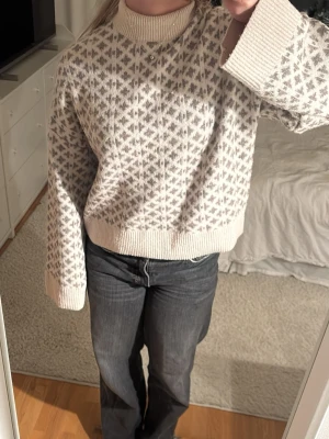Beige och grå stickad tröja med mönster - Mysig stickad tröja i beige med grått geometriskt mönster. Tröjan har hög krage, breda muddar och en lite boxig, loose passform. Perfekt för kyliga dagar och ger en avslappnad men ändå stilren look.