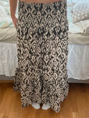 Mönstrad långkjol från Zara - Säljer en svart och beige långkjol från Zara med unikt grafiskt mönster. Kjolen har smock i midjan och volang nertill för en flowy känsla. Perfekt för sommaren! I storlek M men passar S också 