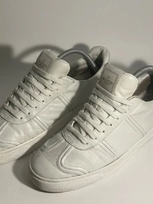 Valentino flycrews - Säljer ett par vita Valentino sneakers i skinn med klassisk låg profil och snörning. Skorna har diskreta sömdetaljer och små nitar baktill på sulan för en edgy touch. Perfekta för dig som gillar stilrena och exklusiva sneakers.Byten kan va intressant‼️