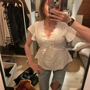 Vit blus med spets och volang - Superfin vit blus med v-ringning, spetsdetaljer och volang vid ärmarna. Blusen har korta ärmar och ett broderat hålmönster som ger en romantisk känsla. Perfekt till jeans eller kjol för en söt look.