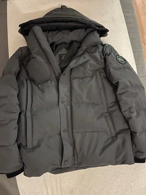 Mörkgrå pufferjacka från Canada Goose - Säljer en mörkgrå pufferjacka från Canada Goose med huva och tydlig logotyp på ärmen. Jackan har dragkedja och knappar framtill, stora fickor och quiltad design. Insidan har praktiska remmar och meshficka. Perfekt för kalla dagar. Köpte den fast den va lite stor så måste få bort den snabbt priset går att diskutera 