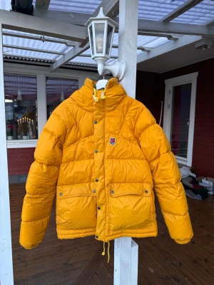 Gul Fjällräven Expedition dunjacka - Säljer en ikonisk gul Fjällräven Expedition Down Jacket med stor huva, två rejäla fickor med knappar och snörning nedtill. Jackan är puffer-modell och har en olivgrön insida. Perfekt för kalla vinterdagar och äventyr i skidbacken! Lite skrynklig på bilderna pga nerpackad sedan förra vintern! Endast använd en halv vinter. Nypris drygt 8000kr.