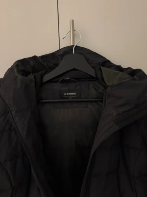 Svart pufferjacka från Everest - Svart pufferjacka från Everest med huva och dragkedja framtill. Jackan har quiltad design och två sidofickor med dragkedja. Perfekt för kalla dagar och har en stilren look som funkar till det mesta.