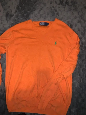Orange tröja från Polo Ralph Lauren - Säljer en orange långärmad tröja från Polo Ralph Lauren med v-ringning och den klassiska gröna loggan broderad på bröstet. Perfekt för dig som vill ha en färgstark och stilren look. Tillverkad i mjuk bomull för skön känsla.