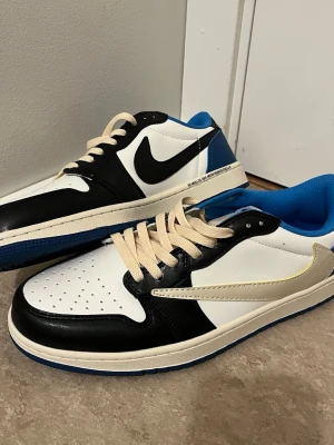 Nike Air Jordan 1 Low Travis Fragment - Nike Air Jordan 1 Low Travis Scott x Fragment med svart, vit och blå färg. Skorna har omvänd vit swoosh, broderade detaljer på hälen och beige snörning. Tillverkade i skinn med blå yttersula och perforerad tå. Perfekt för dig som gillar unika sneakers och collabs.