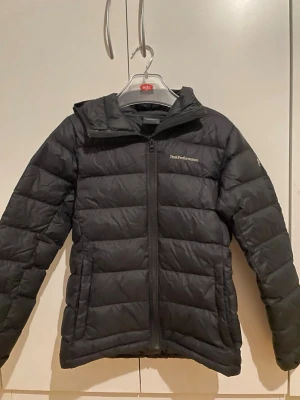 Svart dunjacka Peak Performance - Svart dunjacka från Peak Performance med huva och dragkedja framtill. Jackan har ett quiltat mönster och logga broderad på bröstet. Perfekt för kalla dagar och har en stilren look som funkar till många outfits.