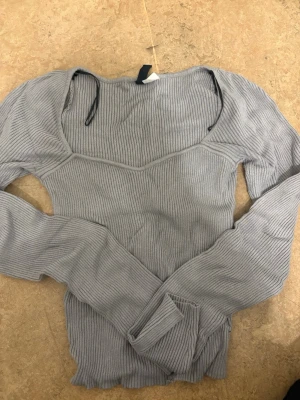 Grå ribbad långärmad topp XS - Säljer en grå ribbad långärmad topp i storlek XS. Toppen har en snygg, figurnära passform med markerad söm över bröstet och lätt vågad kant nedtill. Perfekt att styla med jeans eller kjol för en cool look.