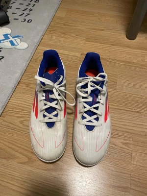 Vita Adidas inomhus fotbollsskor med blå detaljer - Säljer ett par snygga Adidas fotbollsskor i vitt med blå och röda detaljer. Skorna har klassiska tre ränder på sidan, snörning och låg profil. Insidan är blå och ovandelen är tillverkad i syntetmaterial med textur för bättre bollkontroll. Man kan diskutera om priset 