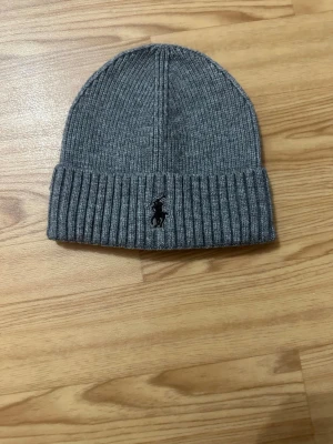Grå ribbstickad mössa Polo Ralph Lauren - Säljer en grå ribbstickad mössa från Polo Ralph Lauren med klassisk broderad logga framtill. Mössan är tillverkad i en mjuk blandning av bomull, polyester och nylon och har vikt kant för extra stilpoäng.