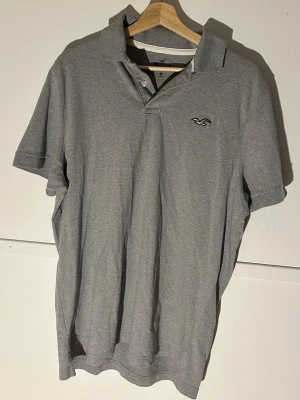 Grå pikétröja från Hollister XL - Säljer en grå pikétröja från Hollister i storlek XL. Tröjan har klassisk krage, knappar vid halsen och Hollisters logga broderad på bröstet. Materialet är mjukt och stretchigt, perfekt för en chill och stilren look.