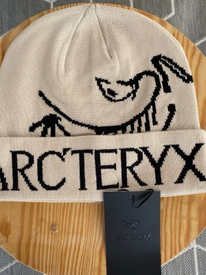 Beige och svart Arc'teryx mössa - Arc’teryx Ascent Beanie – Beige – Aldrig använt!  En stilren och funktionell mössa från Arc’teryx i beige med det klassiska logotrycket. Tillverkad i mjukt och slitstarkt material som håller dig varm både till vardags och vid utomhusaktiviteter. Aldrig använt, taggen sitter fortfarande kvar.  Arc’teryx Ascent Beanie – Beige – Never worn!  A clean and functional beanie from Arc’teryx in beige, featuring the classic logo design. Made from soft and durable material that keeps you warm for everyday 