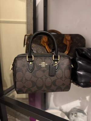 Brun handväska från Coach - Säljer en snygg brun handväska från Coach med klassiskt logomönster och svarta detaljer. Väskan har guldfärgade metalldetaljer, dubbla handtag och dragkedja upptill. Insidan har fack och en avtagbar axelrem. 