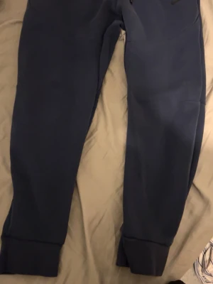 Mörkblå mjukisbyxor från Nike - Säljer ett par mörkblå mjukisbyxor från Nike med smal passform och breda muddar vid bensluten. Byxorna har en enkel design och är tillverkade i mjukt material som är skönt att ha på sig. Perfekta för chill eller träning. Nike tech 