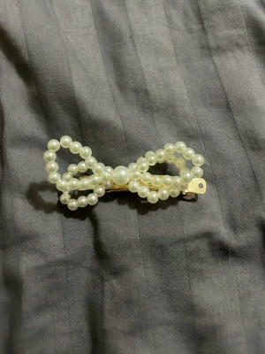 Pärlrosett hårspänne i guld - Säljer ett hårspänne format som en rosett, dekorerat med vita pärlor och med en guldfärgad metallbas. Perfekt för att ge frisyren en söt och elegant touch. Spännet är lätt att använda och håller håret på plats.