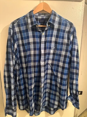 Blårutig skjorta från GANT, storlek L - Säljer en blårutig skjorta från GANT i storlek L. Skjortan har klassisk krage, knappar framtill och en bröstficka med broderad logga. Tillverkad i mjuk bomull och har långärmad passform. Perfekt för dig som gillar stilrena och tidlösa mönster.