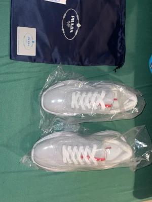 Vita sneakers från Prada - Säljer ett par fräscha vita sneakers från Prada med klassisk röd logga på plösen. Skorna har snörning och är tillverkade i en kombination av syntet och skinn, vilket ger en clean och modern look. Perfekta för dig som gillar exklusiva märken och stilren design.