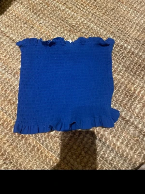 Blå bandeau tubtopp från H&M - Säljer en blå bandeau tubtopp från H&M i storlek M. Toppen är smockad och har volangdetaljer upptill och nedtill för en söt look. Perfekt för varma dagar och enkel att matcha med jeans eller kjol.