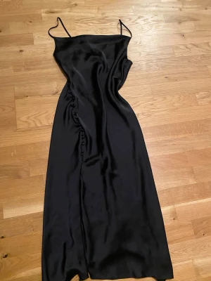 Svart långklänning i satin - Säljer en svart långklänning med tunna axelband och slät, glansig yta. Klänningen har en enkel och elegant siluett som faller snyggt längs kroppen. 