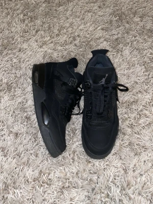 Svarta Nike Jordan 4 sneakers - Säljer ett par svarta Nike Jordan 4 sneakers med cool meshpanel, snörning och Jumpman-logga på plösen. Skorna har en platt sula och detaljer i syntet och skinn. Perfekt för dig som gillar streetstyle och vill ha en klassisk siluett med modern vibe.