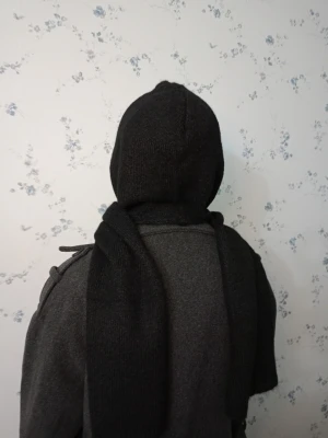 Svart stickad väst med huva - Svart stickad väst med huva och breda axelpartier. Västen har en enkel, rak form och är tillverkad i mjukt stickat material. Perfekt för lager-på-lager och ger en cool vibe till din outfit. Snygg att bära över en långärmad tröja eller jacka.