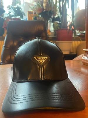 Svart keps från Prada - Snygg svart Prada keps som har används i mindre än en vecka och är i helt nytt skick, den går att justera till alla möjliga storlekar så den passar alla. Kepsen är perfekt för vintern och förvarar värmen runt huvudet.  Kepsen är gjord i skinn och perfekt för dig som gillar exklusiva accessoarer… priset går att pruta.