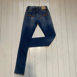Blå jeans från Nudie - Säljer ett par klassiska blå jeans från Nudie med snygga orangea sömmar och broderade bakfickor. Modellen är en skinny modell som har en normal passform och raka ben, tillverkade i slitstarkt bomullsmaterial. Perfekt för dig som gillar stilrena och tidlösa jeans. Storleken på jeansen är W27 L32. Midjan är 33cm. Längden är 104cm och bredden på smalbenet är 15cm