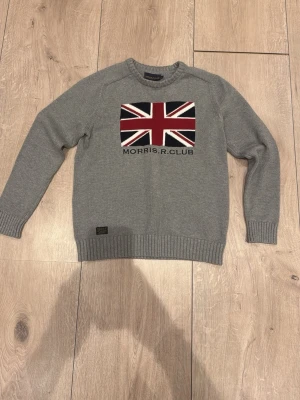 Grå stickad tröja Morris R Club - Snygg grå stickad tröja från Morris R Club med Union Jack-flagga i rött, vitt och blått på bröstet. Tröjan har rund hals, ribbade muddar och lång ärm. Perfekt för dig som gillar klassisk stil med en twist.