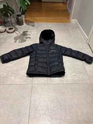 Svart Peak Performance dunjacka - Säljer en svart jacka från Peak Performance (Frost down puffer jacket JR) 2700 nypris. med huva och snyggt broderad logga på bröstet. Jackan har quiltad design, dragkedja framtill och långärmade ärmar. Perfekt för kalla dagar och har en clean, sportig look. Kan disskutera priset