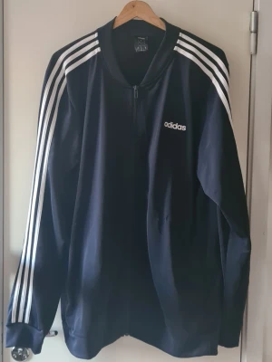 Svart Adidas vindjacka XL - Säljer en svart Adidas vindjacka i storlek XL med klassiska vita ränder längs ärmarna och Adidas-logga på bröstet. Jackan har dragkedja framtill och är långärmad. 