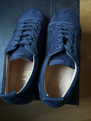 Marinblå mockasneakers från Louboutin - Säljer ett par marinblå sneakers från Christian Louboutin i mjuk mocka med klassisk röd sula och snygg snörning. Skorna har rund tå och diskret broderad logga på hälen. Perfekta för dig som vill ha något exklusivt och stilrent.