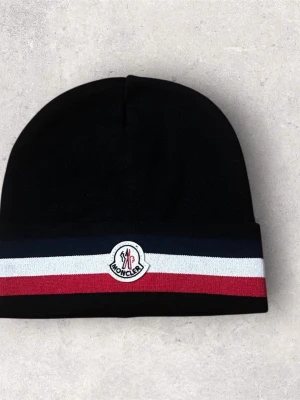 Svart Moncler mössa med ränder - Snygg svart mössa från Moncler med breda ränder i marinblått, vitt och rött längs kanten. Mössan har Moncler-logga framtill och är stickad i mjukt material som håller dig varm. Perfekt för dig som vill ha en stilren accessoar med lyxig känsla.