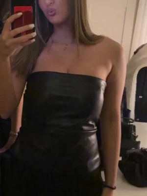 Svart tubtopp i skinnimitation - Säljer en svart axelbandslös tubtopp i skinnimitation. Toppen har en stilren och minimalistisk look med slät yta och dragkedja i sidan för enkel på- och avtagning. Perfekt för dig som vill ha en edgy vibe till din outfit.