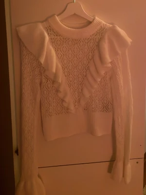 Vit stickad tröja med volanger - Säljer en vit stickad tröja med snygga volangdetaljer längs axlarna och ner mot bröstet. Tröjan har ett genombrutet mönster och långa ärmar med ribbade muddar. Perfekt för dig som gillar romantisk och trendig stil.