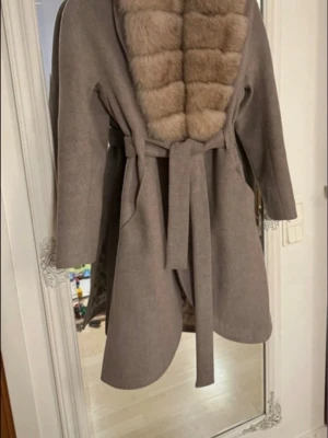 Beige kappa med päls och bälte str 36 - Säljer en beige kappa med bred, fluffig äkta päls i ljusbrun ton längs kragen och bröstet. Jackan har ett matchande bälte i midjan och quiltat foder på insidan. Perfekt för dig som vill ha en lyxig och varm look under höst och vinter.