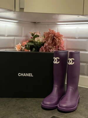 Lila stövlar från Chanel - Säljer ett par lila stövlar från Chanel med vit logga framtill och präglad Chanel-text på hälen. Stövlarna är gjorda i syntetmaterial och har en platt sula med mönstrad undersida och Chanel-logga. Perfekt för dig som vill sticka ut med en lyxig och färgstark look.