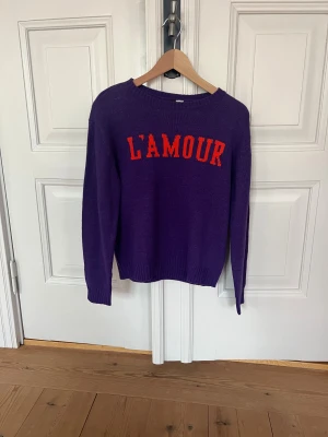 Lila stickad tröja L'AMOUR från Divided - Cool lila stickad tröja från Divided/HM med röd text 'L'AMOUR' = kärleken. Materialet är tunt stickad akryl. Hylla kärleken med detta statement plagg!